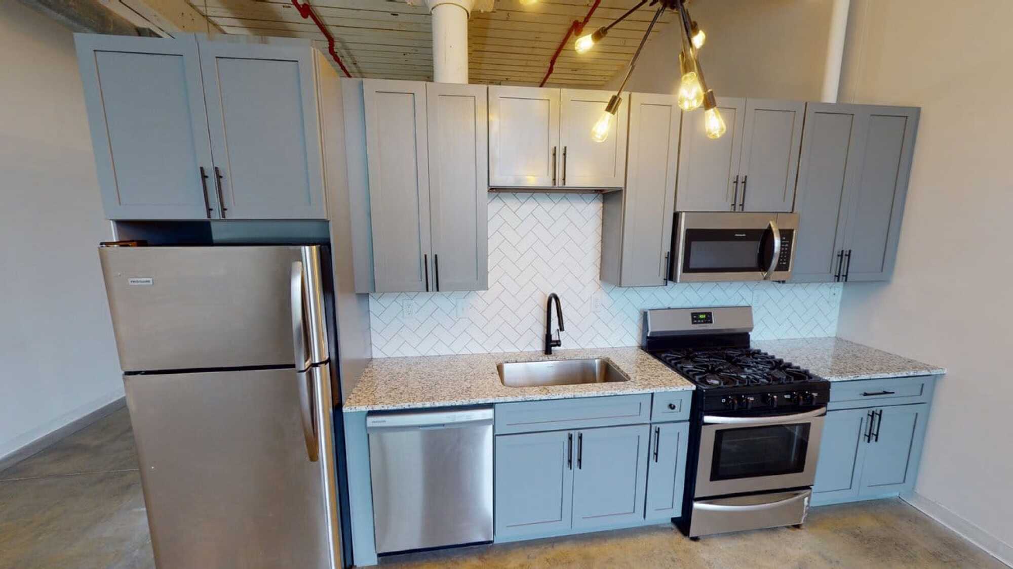 Modern kitchen with gray cabinets, stainless steel appliances, herringbone tile backsplash, and exposed ceiling pipes.