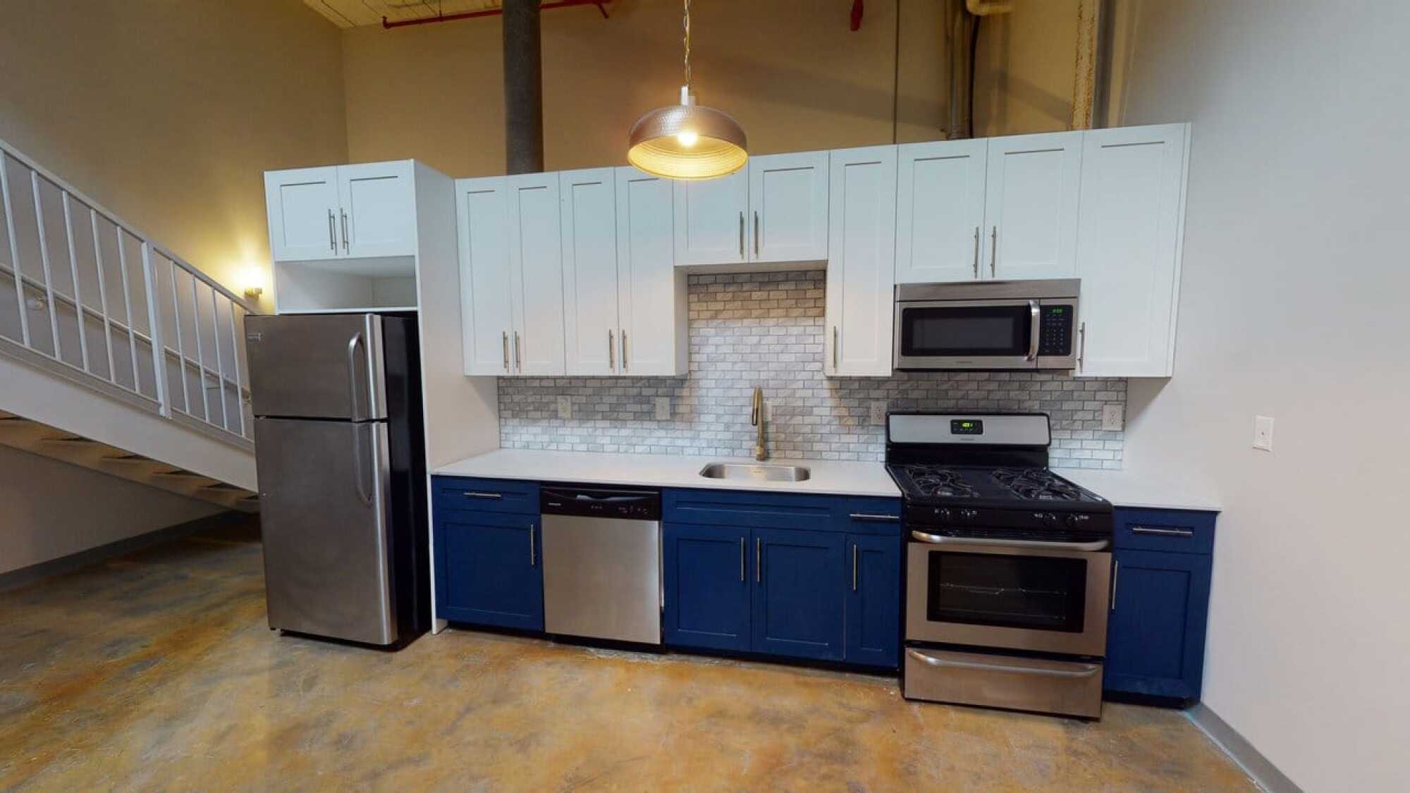 Modern kitchen with white upper and blue lower cabinets, stainless steel appliances, and tile backsplash under a hanging light.