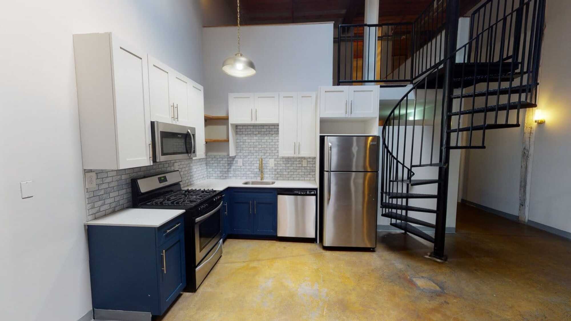 Modern kitchen with blue and white cabinets, stainless steel appliances, and a spiral staircase nearby.