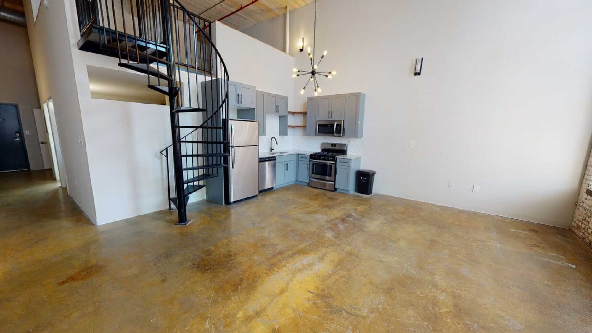 Modern loft kitchen with gray cabinets, stainless steel appliances, spiral staircase, and polished concrete floor.