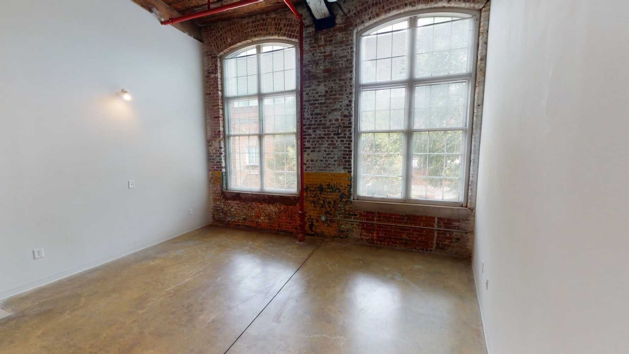 Empty room with brick walls, two large arched windows, concrete floor, and a single wall-mounted light fixture.