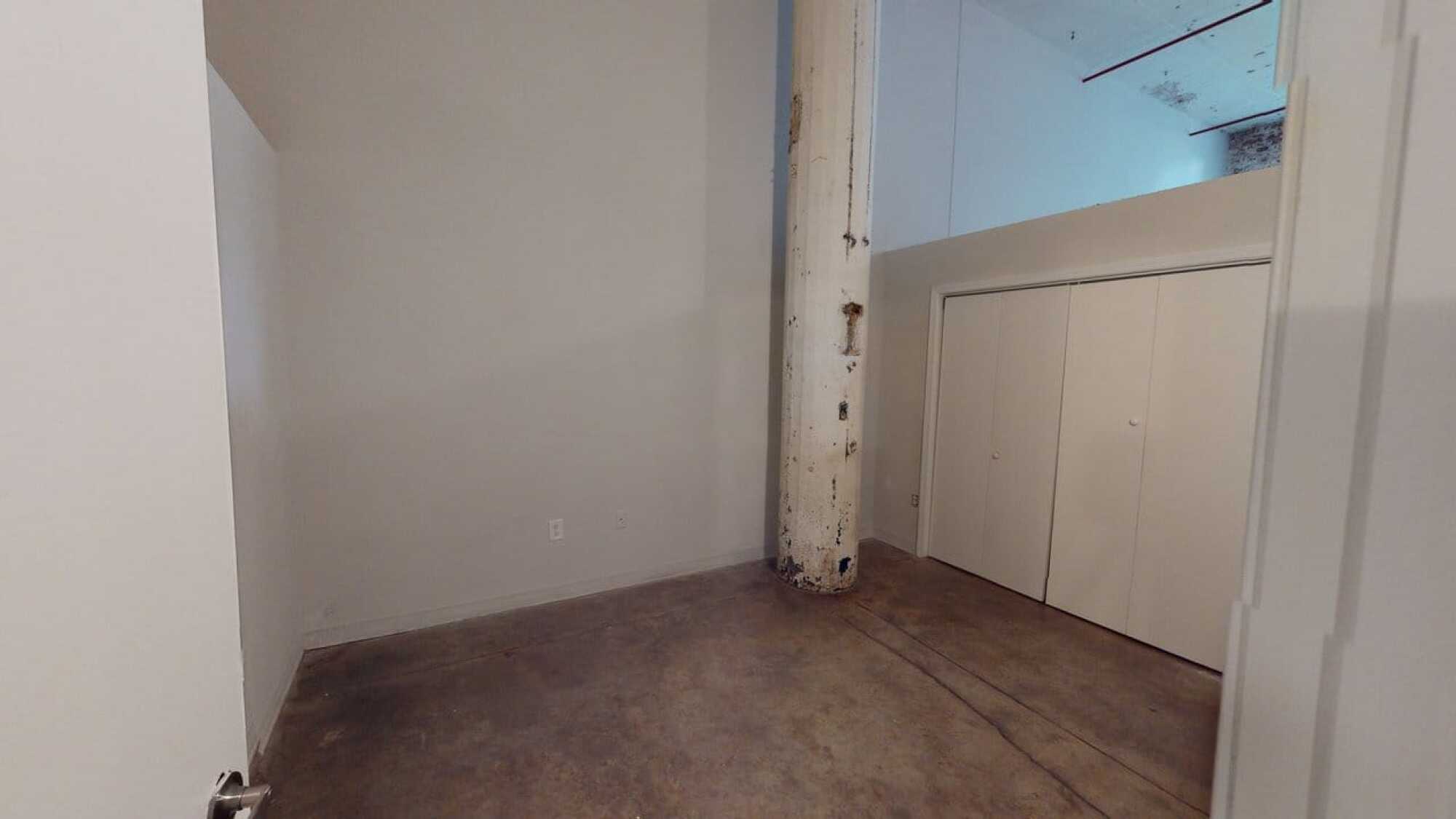 Empty room with concrete floor, white walls, a large column, and closed closet doors beneath a lofted area.