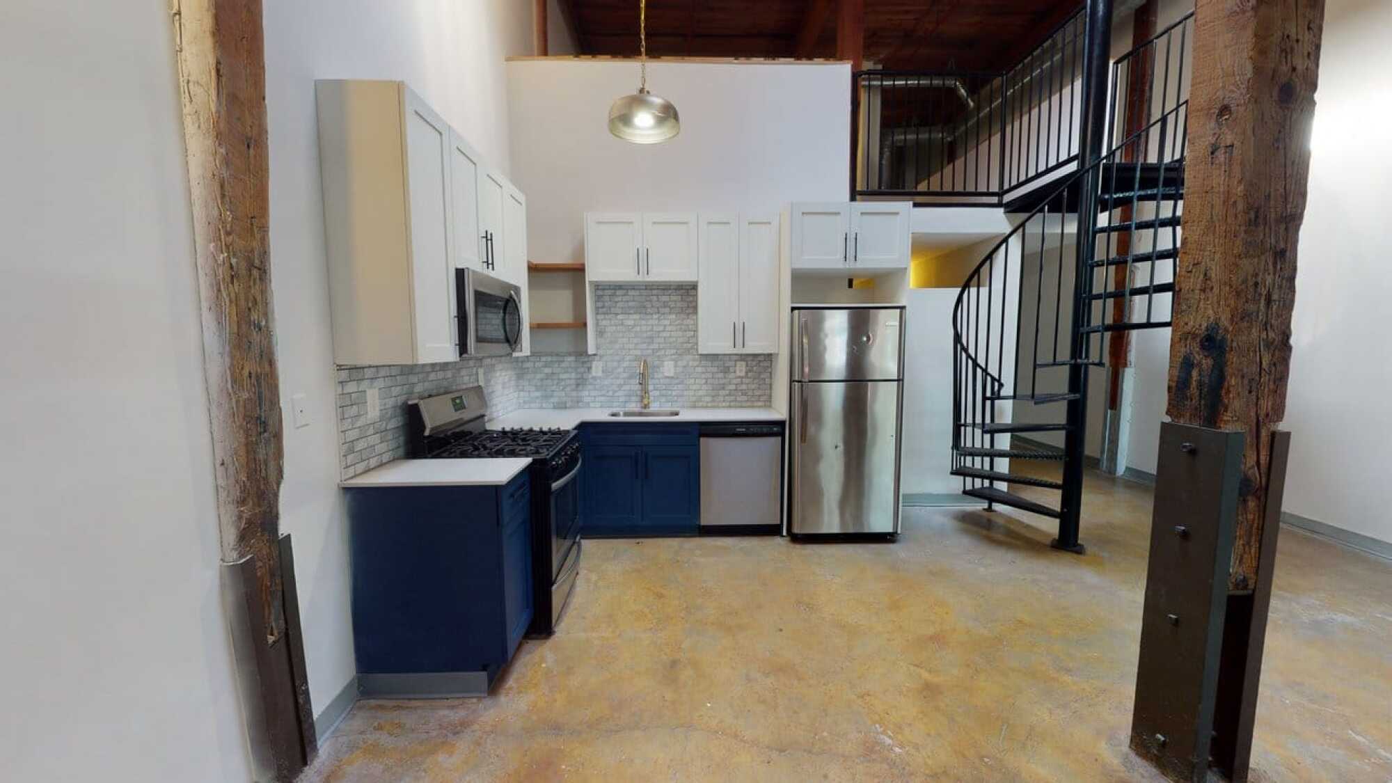 Loft kitchen with blue and white cabinets, stainless appliances, and a spiral staircase leading to an upper level.