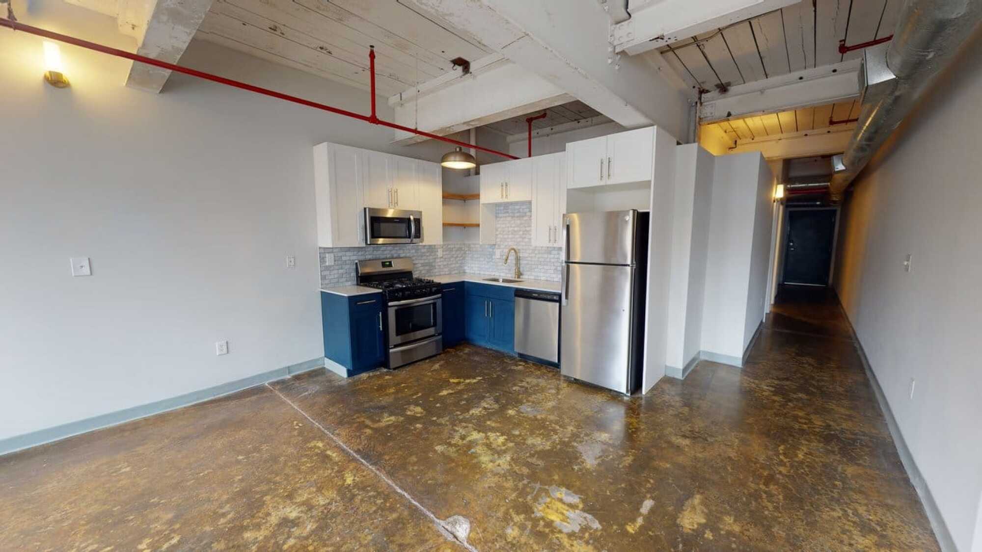 Modern kitchen with blue and white cabinets, stainless steel appliances, and exposed pipes in a loft-style apartment with concrete floors.