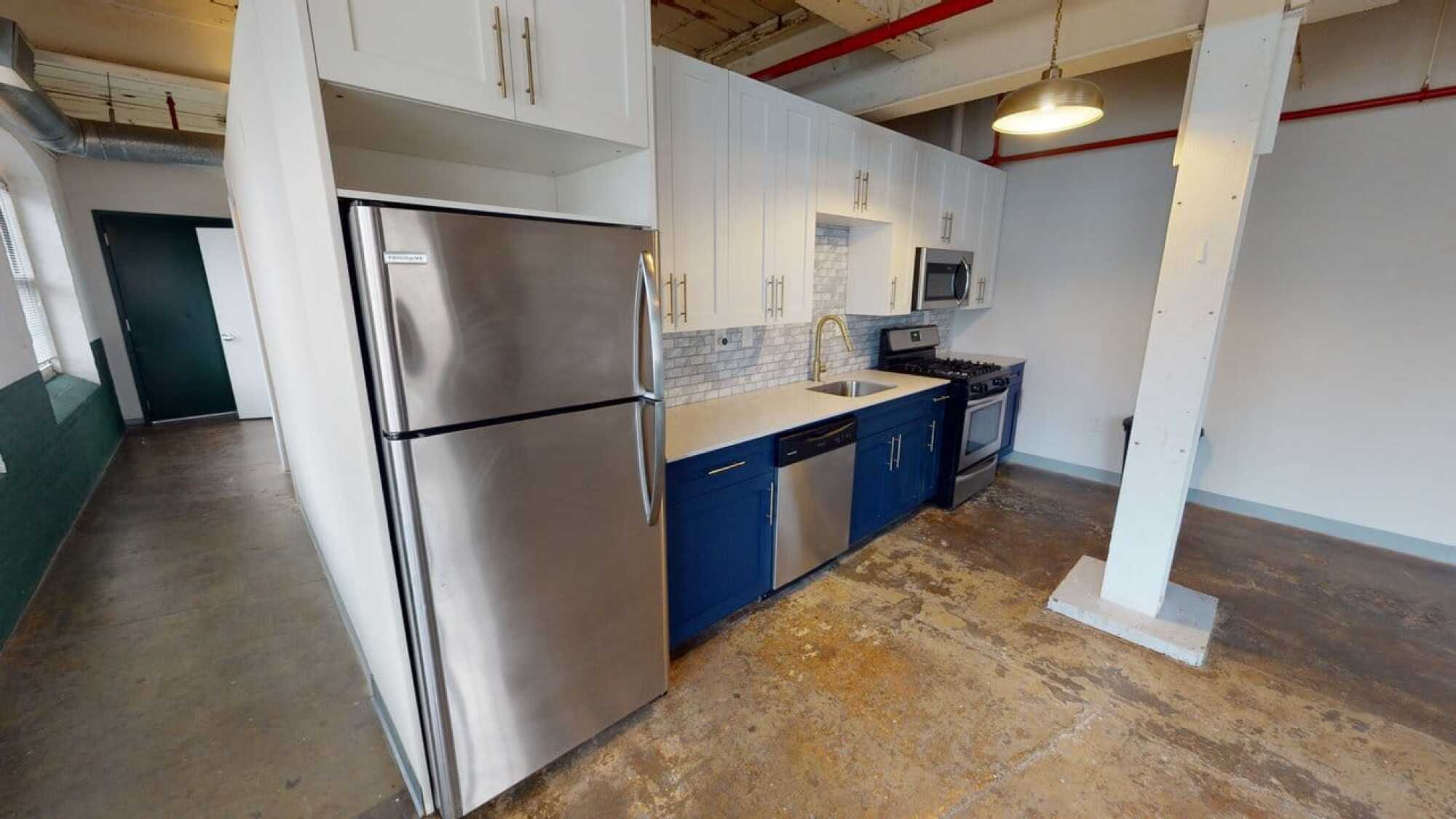 Modern kitchen with stainless steel fridge, white cabinets, blue lower cabinets, stove, microwave, and exposed ceiling pipes.