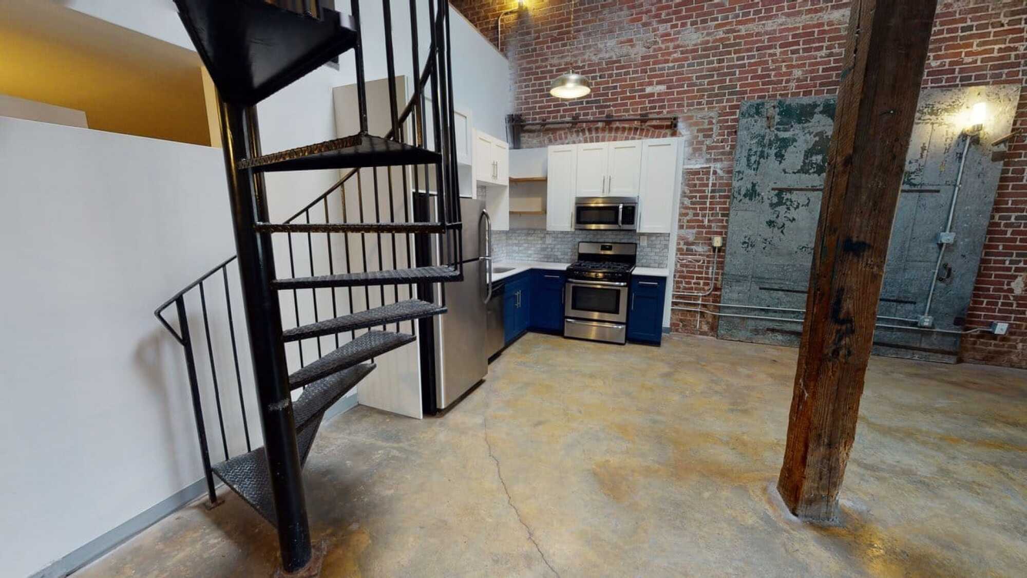 Modern loft kitchen with brick walls, spiral staircase, blue and white cabinets, and exposed beams.
