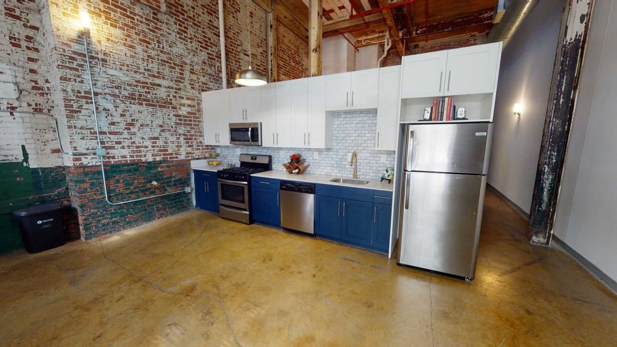 Modern loft kitchen with exposed brick, stainless steel appliances, blue lower cabinets, and white upper cabinets.