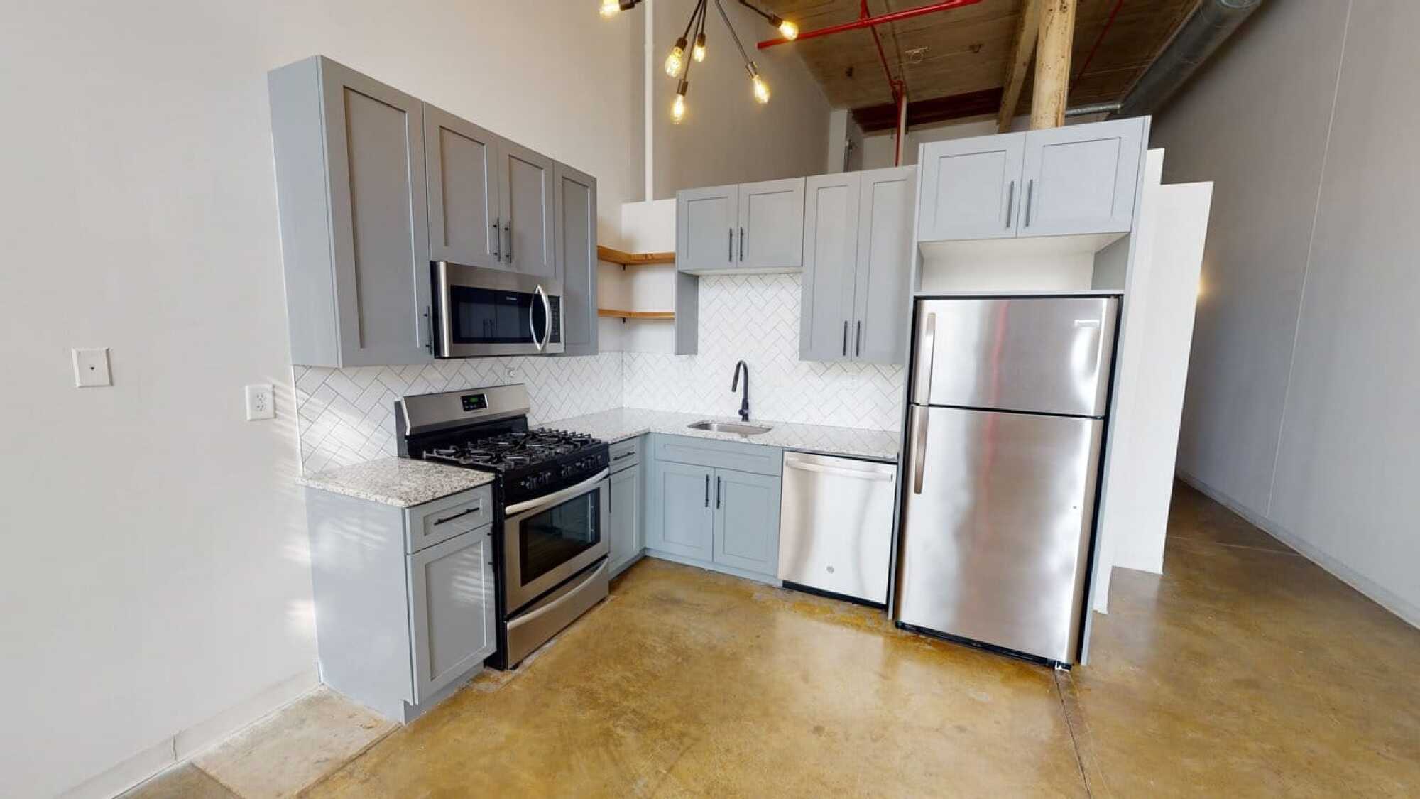 Modern kitchen with gray cabinets, stainless steel appliances, white tile backsplash, and exposed ceiling pipes.