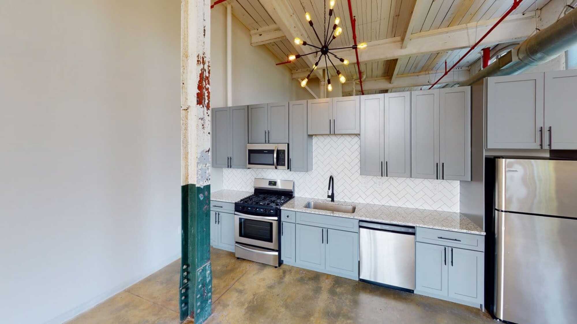 Modern kitchen with gray cabinets, stainless steel appliances, unique ceiling light, and exposed pipe and beam elements.