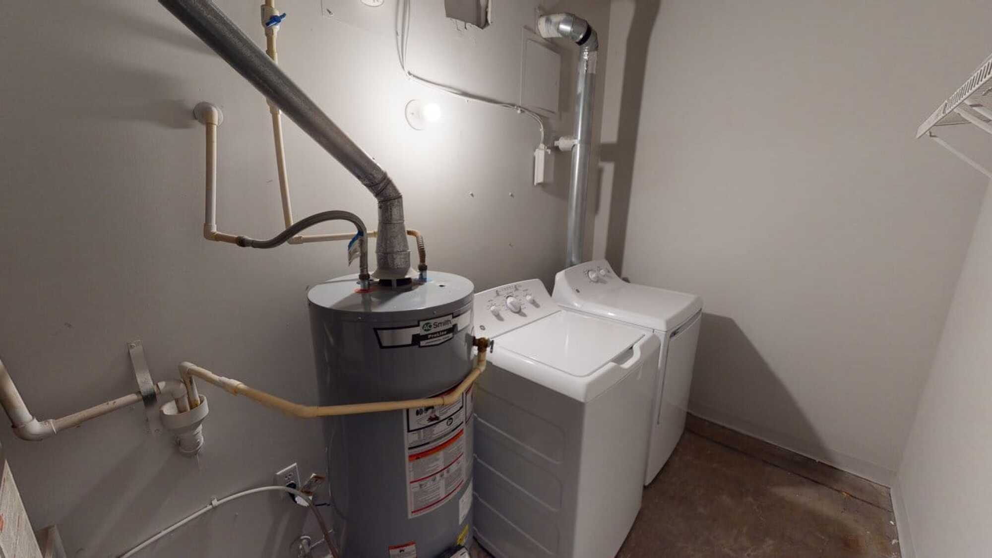 A water heater and a washer and dryer are in a small utility room with exposed pipes and a bare floor.