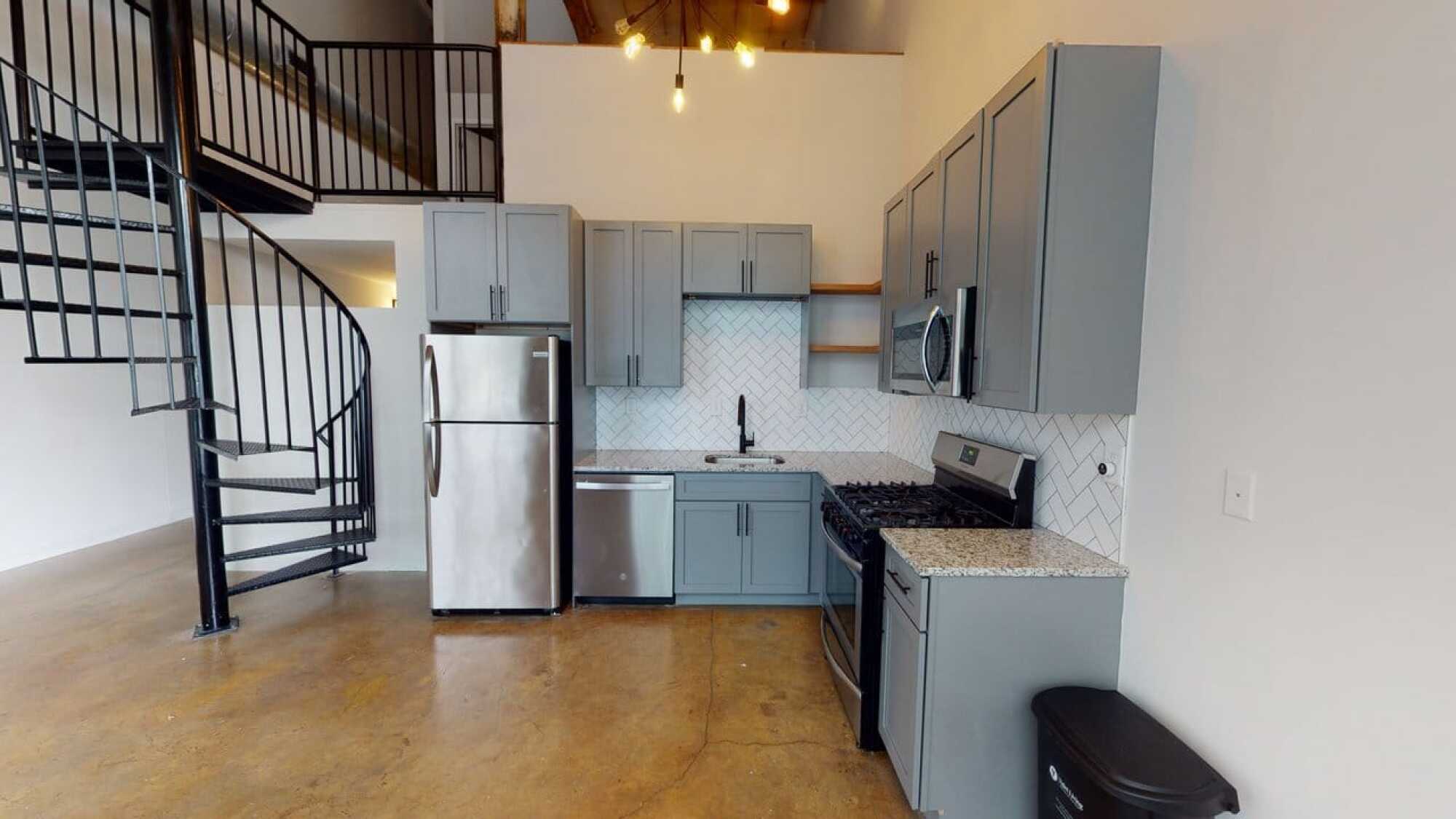 Modern kitchen with gray cabinets, stainless steel appliances, and a spiral staircase on a polished concrete floor.
