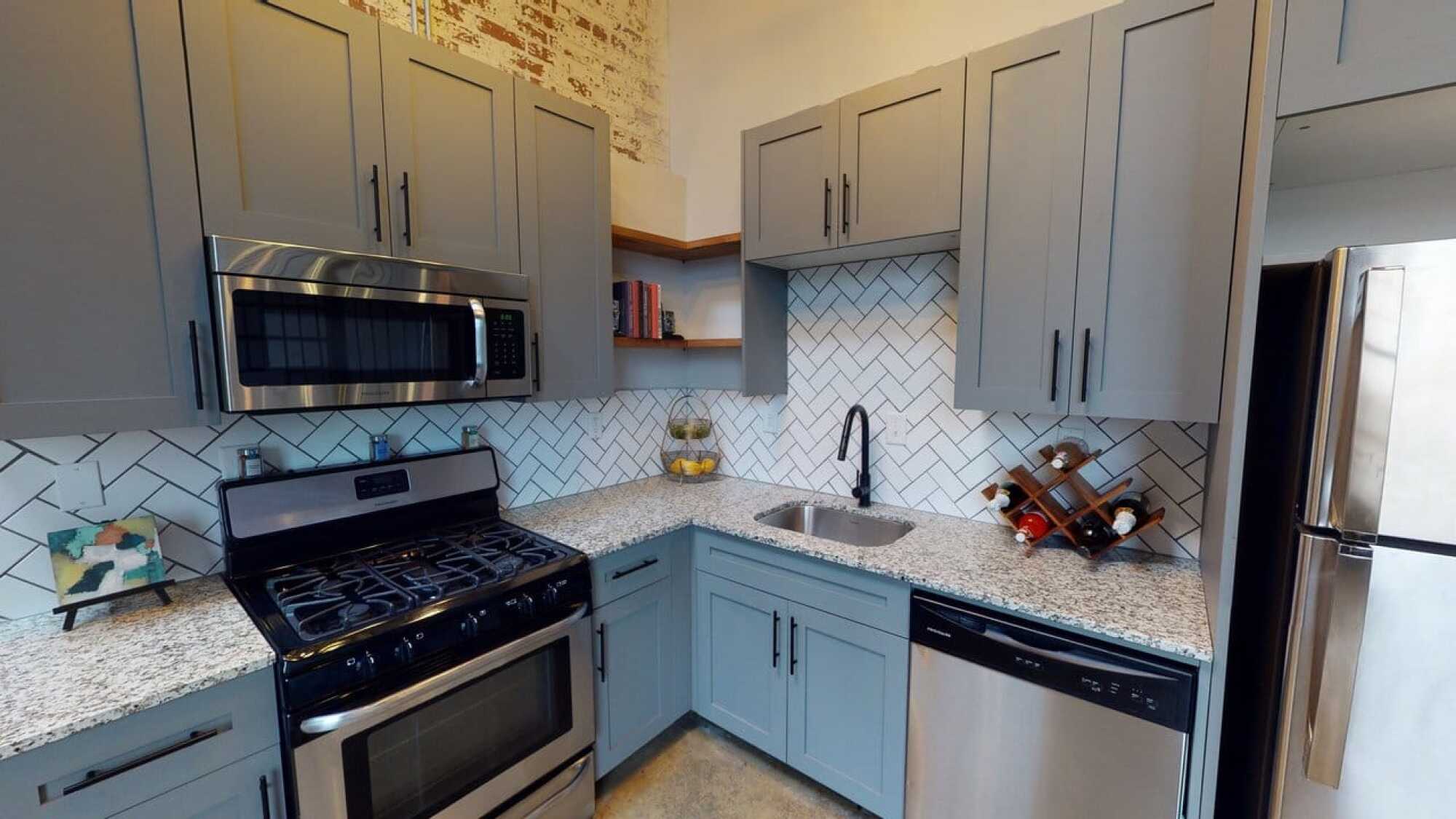 Modern kitchen with gray cabinets, stainless steel appliances, granite countertops, and white herringbone tile backsplash.