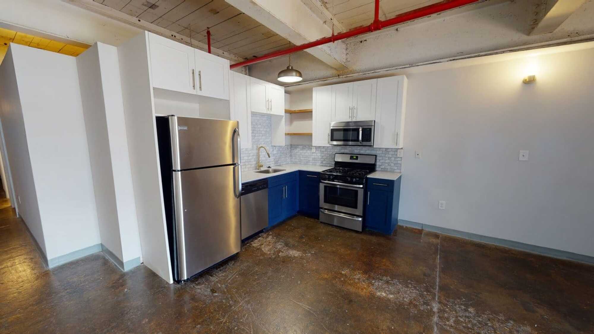 Modern kitchen with stainless steel appliances, white and blue cabinets, tile backsplash, and exposed ceiling pipes.