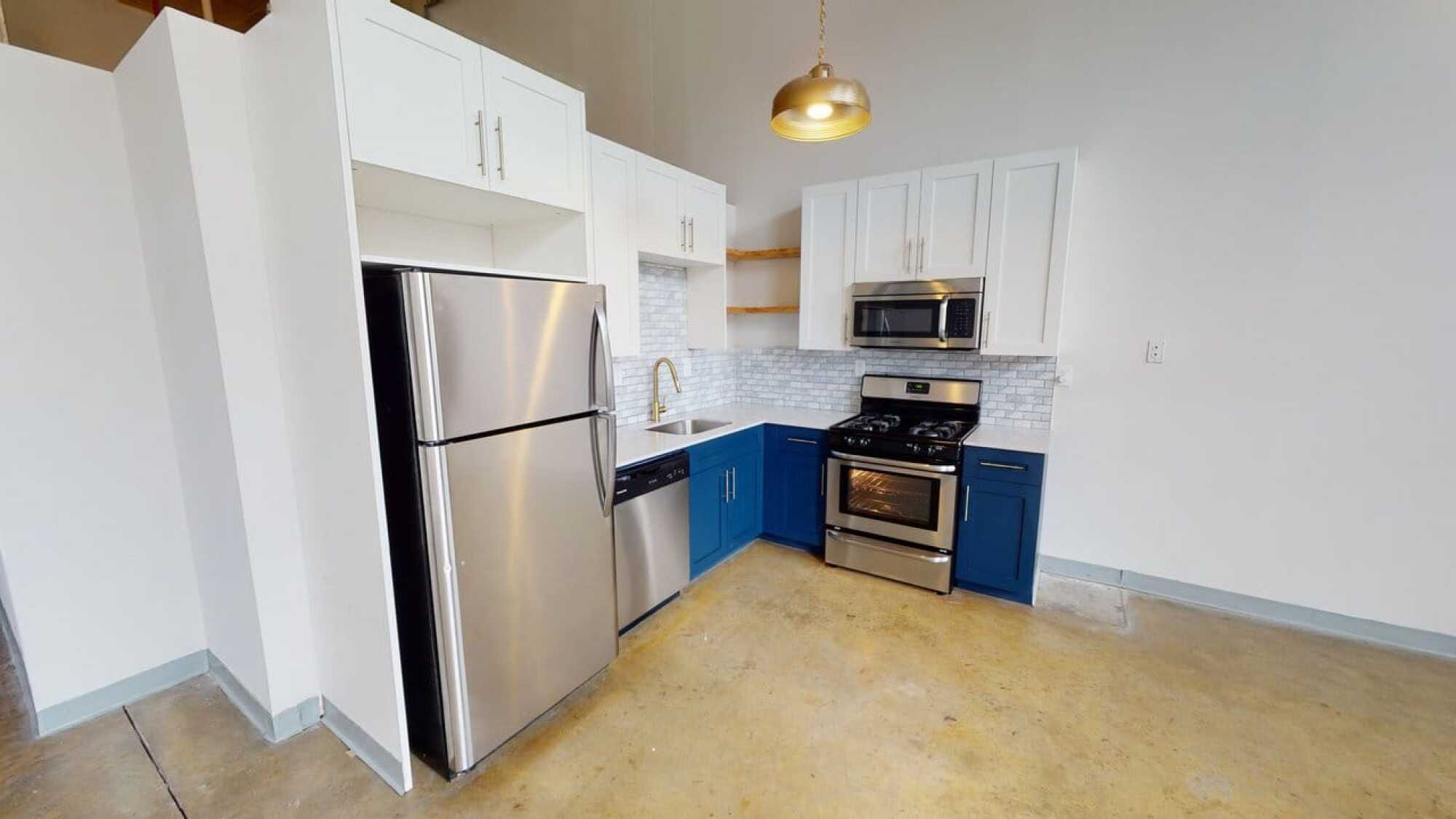 Modern kitchen with stainless steel appliances, blue and white cabinets, gold faucet, and pendant light over concrete floor.