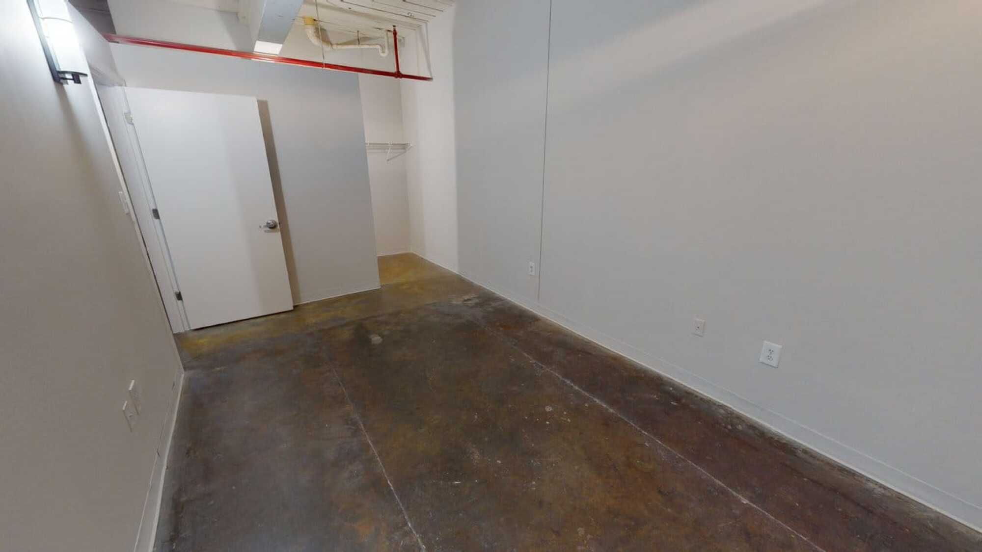 Empty, narrow room with concrete floor, white walls, a closet, and door open to the left. Red pipe runs along the ceiling.