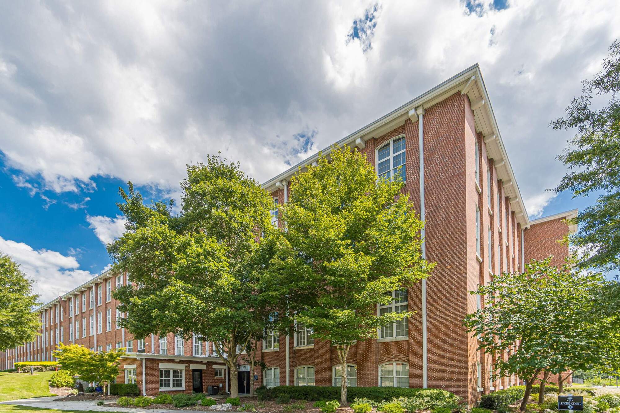 Red-brick apartment building with large windows, trees in front, and partly cloudy sky above.